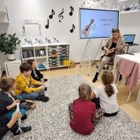 Na zdjęciu widzimy nauczycielkę grającą na ukulele, otoczoną dziećmi w sali edukacyjnej. Dzieci słuchają lub bawią się instrumentami. W tle tablica interaktywna z tekstem i dekoracje w muzyczne nuty.