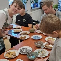 Zdjęcie przedstawia uczestników dekorujących świąteczne pierniki podczas warsztatów kulinarnych. Na stołach leżą talerze z ciastkami i kolorowymi posypkami, a dzieci i dorośli pracują z zapałem, tworząc piękne ozdoby z lukru.