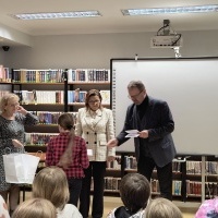 Mężczyzna wręcza dyplom dziewczynce w czerwonej koszuli w bibliotece. Obok stoją dwie kobiety — jedna trzyma torbę z upominkiem. W tle widoczne są półki z książkami i tablica multimedialna.