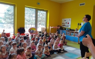 Na zdjęciu widać grupę dzieci siedzącą na kolorwym dywanie, w tle żółta ściana i duże okna. 