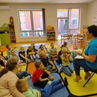 Kobieta w niebieskiej bluzce siedzi na krześle i pokazuje dzieciom otwartą książkę. Przedszkolaki siedzą w półkolu i słuchają z zaciekawieniem. W tle kolorowe szafki, okna i pomoce dydaktyczne.
