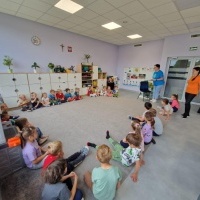 Dzieci siedzą w kręgu na dywanie w sali przedszkolnej. W pomieszczeniu znajdują się szafki, zabawki, plansze edukacyjne i dwie osoby dorosłe stojące przy drzwiach.