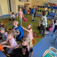 Na zdjęciu widać grupę dzieci w sali. Część siedzi na kolorowym dywanie, inne stoją lub chodzą. Na stolikach leżą książki. W tle są półki z zabawkami i materiałami edukacyjnymi.
