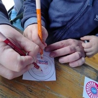Na zdjęciu widoczne ręce jednego dorosłego oraz jednego dziecka malujące coś na kartce za pomocą kredek.