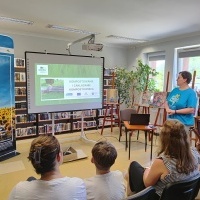 Na zdjęciu widać prezentację pt. „Kompostowanie i zakładanie kompostownika” wyświetlaną na ekranie w bibliotece. Przed ekranem siedzą osoby, obok stoi kobieta w niebieskiej koszulce. W tle półki z książkami i obrazy.