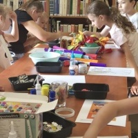 Dzieci biorą udział w zajęciach plastycznych w bibliotece - rysują, kolorują i tworzą prace z materiałów dekoracyjnych przy stole pełnym przyborów.
