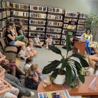 Na zdjęciu widać spotkanie czytelnicze w bibliotece. Kobieta czyta książkę dzieciom, które siedzą z opiekunami. W tle regały z książkami i wystawa lalek. Pomieszczenie jest jasne, z dużymi oknami i rośliną.