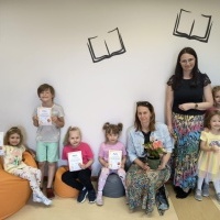 Na zdjęciu widać siedmioro dzieci trzymających dyplomy oraz dwie kobiety - jedna z nich trzyma bukiet kwiatów. W tle biała ściana z czarnymi grafikami otwartych książek.