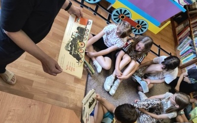 Na zdjęciu widać grupę dzieci siedzących na dywanie w bibliotece, słuchających osoby dorosłej czytającej książkę o lokomotywach. W tle widoczna jest kolorowa dekoracja w kształcie pociągu z książkami.