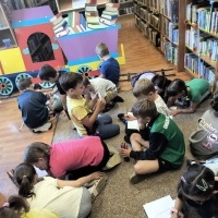 Na zdjęciu dzieci siedzą na dywanie w bibliotece i rysują lub kolorują ilustracje. W tle widać kolorową dekorację w kształcie lokomotywy oraz regały z książkami. Panuje twórcza i radosna atmosfera.