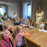 Na zdjęciu widoczna jest grupa dzieci siedzących przy stole oraz trzy kobiety, z których jedna częstuje dzieci cukierkami. W tle widać dwa okna.