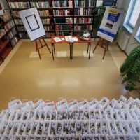Pod ścianą widoczne regały z książkami, oraz stojące dwie sztalugi z plakatami informujących o akcji Przeczytam 52 książki w 2020 roku, oraz stolik z nagrodami i kulą do losowania nagród. Na pierwszym planie widoczna duża ilość torebek papierowych.