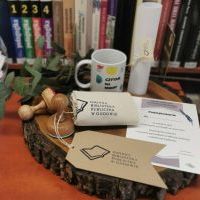 W centralnym miejscu leży plaster drewna, na którym stoi kubek w kolorowe kółka, torba lniana z nadrukowanym logiem biblioteki, dwa drewniane stemple, zielona gałązka, wzór dyplomu w formacie A5, oraz zwinięty rulon kartki papieru z przepasanym logiem.