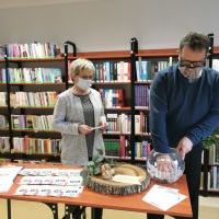 Zdjęcie przedstawiające stojące przed stołem dwie postacie, tj. kobiety i mężczyzny, który pochyla się nad szklaną kulą i losuje. W tle znajdują się regały z książkami.