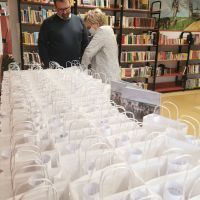 Zdjęcie przedstawiające bardzo dużą ilość torebek papierowych, przy których stoją dwie postacie kobiety i mężczyzny. W tle widać regał z ksiażkami.