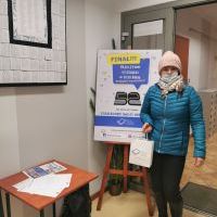 Na zdjęciu widoczna kobieta w niebieskiej kurtce, różowej czapce trzymająca białą torebkę papierową w ręku. 