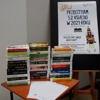 Zdjęcie przedstawiające trzy stosy nowości książkowych wydanych w 2021 roku. Z prawej strony stoi sztaluga na której jest powieszony plakat informacyjny o akcji Przeczytam 52 książki w 2021 roku.