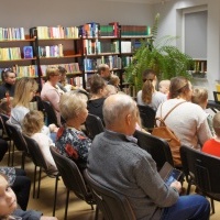 Na zdjęciu grupa osób siedząca na krzesłach w pomieszczeniu bibliotecznym.