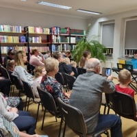 Na zdjęciu grupa osób siedząca na krzesłach w pomieszczeniu bibliotecznym oraz chłopiec grający na organkach.