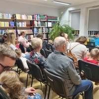 Na zdjęciu grupa osób siedząca na krzesłach w pomieszczeniu bibliotecznym oraz chłopiec grający na organkach.