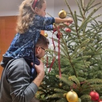 Na zdjęciu widoczna mężczyzna, podnoszący małą dziewczynkę która ubiera świąteczną choinkę. 