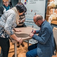 Kobieta pochylająca się nad stolikiem, przy którym siedzi mężczyzna i podpisuje książkę.