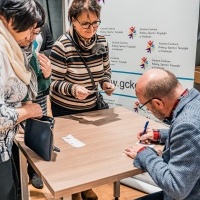 Widoczny mężczyzna podpisujący książkę przy stoliku. Obok stojące dwie kobiety i oczekujące na autograf.