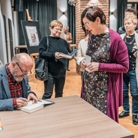 Widoczny mężczyzna podpisujący książkę przy stoliku. Obok kolejka kobiet oczekujących na autograf.