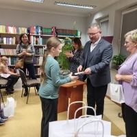 Dziewczynka odbierająca podziękowanie od pani dyrektor. W tle widoczny regał z książkami oraz dwie kobiety i mężczyzna.