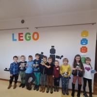 Grupa dzieci trzymajaca w rękach konstrukcje wykonane z klocków Lego. Na ścianie widoczny napis o treści LEGO.