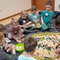 Grupa dzieci siedząca na czarnej folii. Na srodku znajduje się wysypany piasek z kolorowymi kawałkami bibuły.