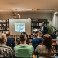 Na zdjęciu widoczna tablica multimedialna oraz grupa ludzi siedzących na krzesłach.