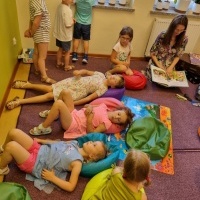 Grupka dzieci leżaca na dywanie. Obok czytająca pani bibliotekarka.