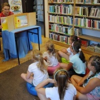 Dzieci siedzące na dywanie i słuchające teatrzyku, które przedstawia i opowiada pani bibliotekarka.