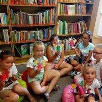 Dzieci siedzące na dywanie i prezentujące własnoręcznie wykonane motyle. W tle regał z ksiażkami.