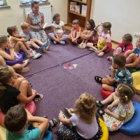 Dzieci siedzące na dywanie w kole i słuchające opowiadania czytanego przez panią bibliotekarkę.