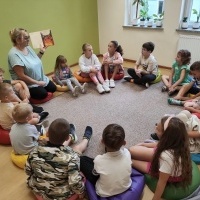 Grupa dzieci siedząca na dywanie wokół pani bibliotekarki, która im czyta.