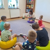 Dzieci siedzące na dywanie i słuchające opowiadnia, które czyta pani bibliotekarka.