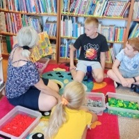 Pani bibliotekarka siedząca wraz z dziećmi na dywanie i czytająca książkę dla dzieci. W tle widoczny regał z książkami.