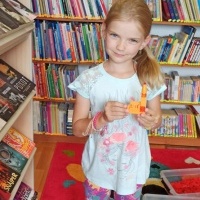 Na tle regału z książkami widoczna dziewczynka z budowlą wykonana z klocków Lego